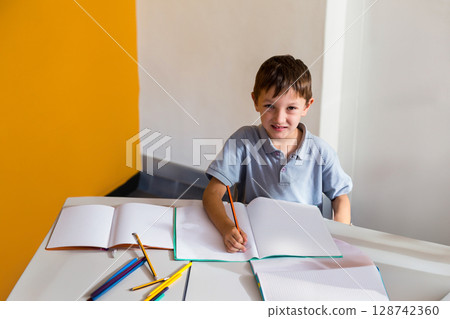 child boy smiling and writing with orange pencil in indoor study area, with scattered notebooks child boy smiling and writing with orange pencil in indoor study area, with scattered notebooks 128742360