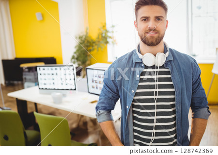 Man in mid 20s standing in modern office, with headphones, monitors and coffee mug, copy space Man in mid 20s standing in modern office, with headphones, monitors and coffee mug, copy space 128742369