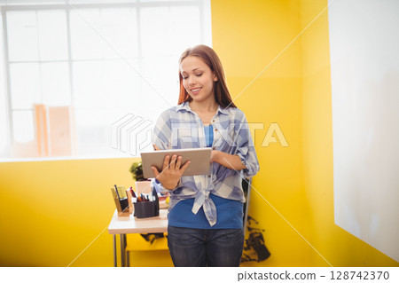 Teenage girl standing and using tablet against yellow wall, with whiteboard and desk plant 128742370