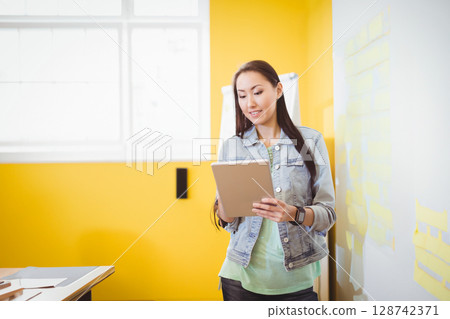 Asian woman standing near center holding tablet in bright creative office, with sticky notes Asian woman standing near center holding tablet in bright creative office, with sticky notes 128742371