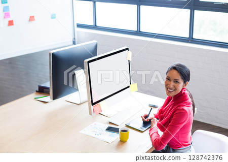 Young Chinese woman creating art on drawing tablet in office, with dual monitors and sticky notes Young Chinese woman creating art on drawing tablet in office, with dual monitors and sticky notes 128742376