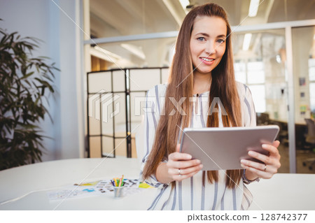 Woman holding tablet and smiling in modern open-plan office, with loose papers and pencil cup 128742377