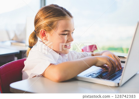 Young girl typing on silver laptop at school desk in modern classroom, with red chair behind Young girl typing on silver laptop at school desk in modern classroom, with red chair behind 128742383