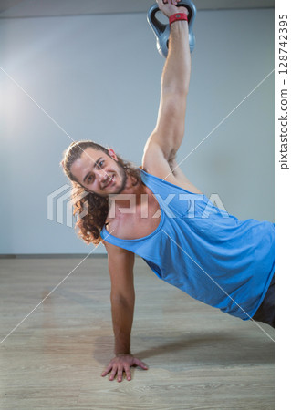 Young man holding kettlebell overhead while balancing in side plank on gym floor with red strap 128742395