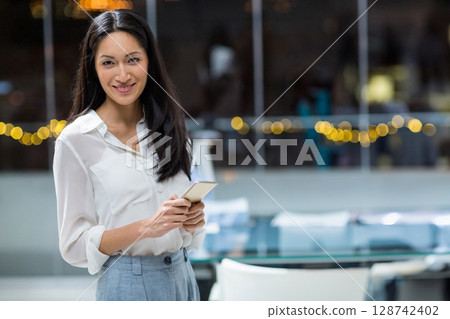 Asian businesswoman standing in office at night, holding smartphone under string lights, copy space 128742402