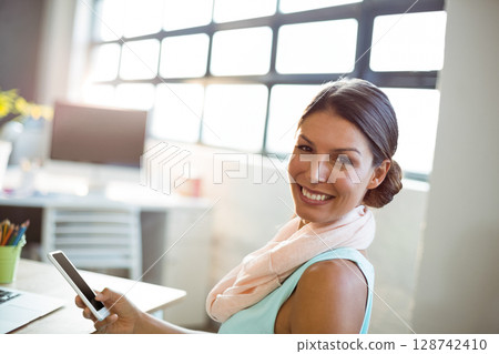 Woman sitting at desk in bright modern office, holding smartphone with laptop and plant, copy space 128742410