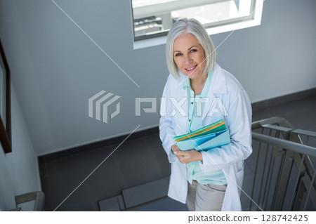 Senior woman in lab coat standing on stairwell landing, holding colored file folders, copy space 128742425