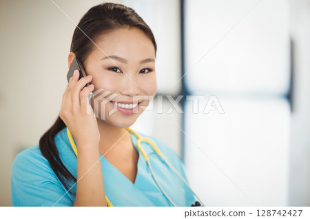 Asian female nurse talking on smartphone in clinic, with yellow stethoscope and bright window 128742427