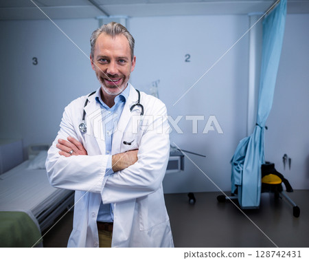 Middle-aged male doctor standing smiling in hospital exam bay, showing stethoscope and IV stand Middle-aged male doctor standing smiling in hospital exam bay, showing stethoscope and IV stand 128742431