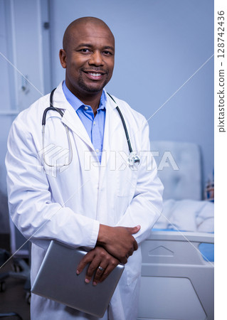 Middle-aged African American man standing in hospital room near bed, holding laptop and stethoscope 128742436