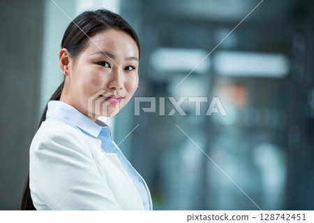 Asian woman standing in modern office with white blazer and blue shirt, smiling, copy space 128742451