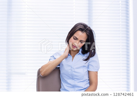 Indian woman rubbing neck sitting on office chair by window with white blinds, feeling discomfort 128742461