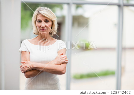 Middle-aged woman standing with arms crossed in room by large windows, overlooking garden outside Middle-aged woman standing with arms crossed in room by large windows, overlooking garden outside 128742462