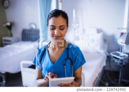 Young Indian nurse wearing stethoscope and using tablet while standing in hospital patient room 128742492