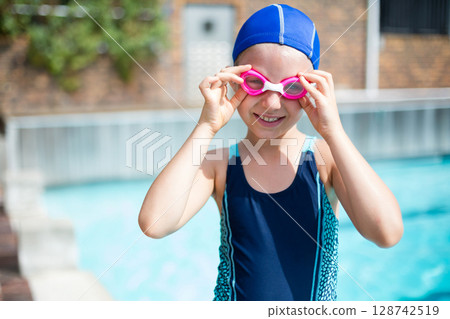 Young girl pulling pink swim goggles over eyes at outdoor pool, wearing blue cap and swimsuit 128742519