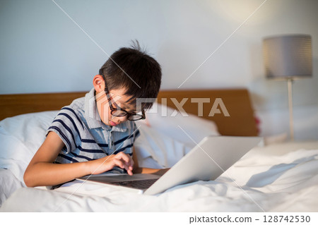 Asian boy child sitting on bed using silver laptop in bedroom with eyeglasses and lamp 128742530