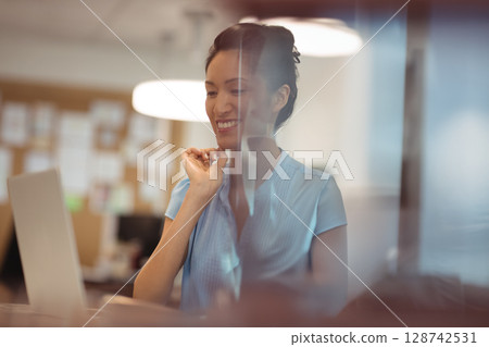 Asian woman typing on silver laptop behind glass partition in office, reflecting pendant lights 128742531