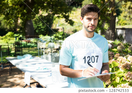 Mid-adult man wearing race bib 1207 standing by folding table in leafy park writing on clipboard 128742533