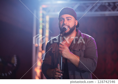 Man gripping mic stand and singing on concert stage, with truss lights and blurred guitar headstock Man gripping mic stand and singing on concert stage, with truss lights and blurred guitar headstock 128742546