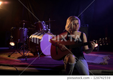 Teenage girl playing acoustic guitar on small indoor stage, with drum kit and amplifier, copy space 128742559