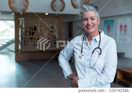Senior female doctor standing in lobby with stethoscope, hanging lights and posters, copy space Senior female doctor standing in lobby with stethoscope, hanging lights and posters, copy space 128742566