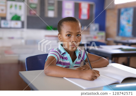 African American child writing with sharpened pencil at desk in sunlit classroom with open notebook African American child writing with sharpened pencil at desk in sunlit classroom with open notebook 128742571