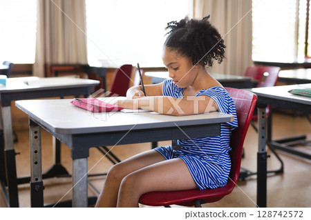7-year-old girl writing on paper at gray desk in classroom, holding pencil and pink pencil case 128742572