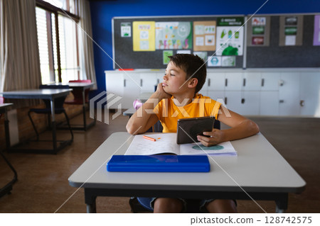 Young boy sitting at desk in classroom, holding tablet with open notebook and pencil box Young boy sitting at desk in classroom, holding tablet with open notebook and pencil box 128742575