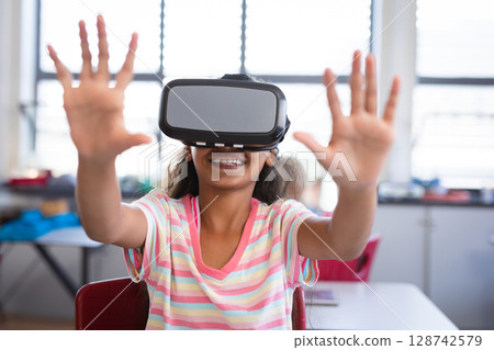 Young girl interacting with virtual environment at classroom desk, wearing virtual reality headset 128742579