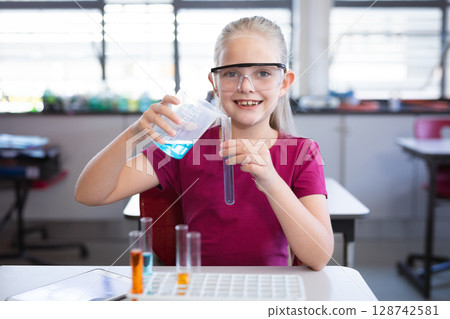 Schoolgirl pouring blue liquid from beaker into test tube at classroom desk with safety goggles Schoolgirl pouring blue liquid from beaker into test tube at classroom desk with safety goggles 128742581