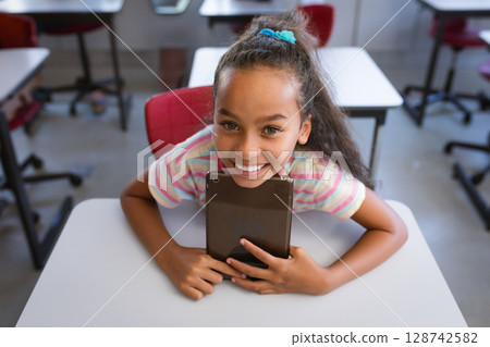 Female child leaning forward on white desk in elementary classroom with red chairs, holding tablet Female child leaning forward on white desk in elementary classroom with red chairs, holding tablet 128742582