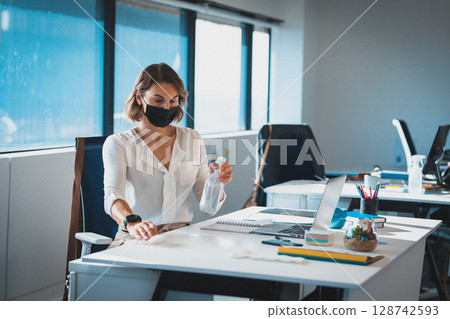 Office worker cleaning desk in open office, with spray bottle and cloth near laptop terrarium 128742593