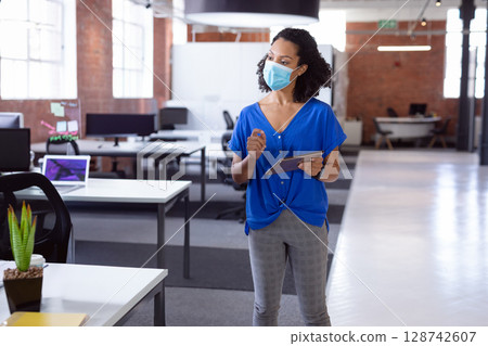Woman wearing surgical mask using tablet and stylus in open-plan office with potted succulent 128742607