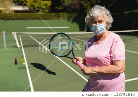 Senior woman standing on tennis court holding racket wearing mask with balls by net, copy space 128742610