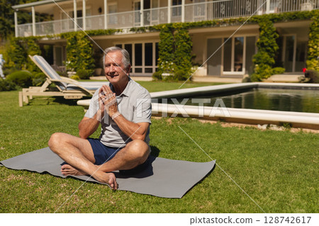 Senior man sitting cross-legged on gray yoga mat in backyard, meditating near pool Senior man sitting cross-legged on gray yoga mat in backyard, meditating near pool 128742617