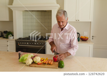 Senior man slicing red bell pepper on wooden cutting board in modern kitchen, with fresh vegetables Senior man slicing red bell pepper on wooden cutting board in modern kitchen, with fresh vegetables 128742620