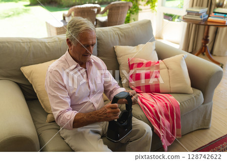 Senior man sitting on beige couch in sunlit living room, using VR headset on charging stand Senior man sitting on beige couch in sunlit living room, using VR headset on charging stand 128742622
