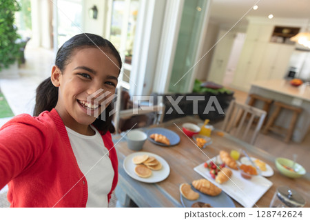 Girl taking selfie with smartphone in open-plan kitchen with wooden breakfast table spread 128742624