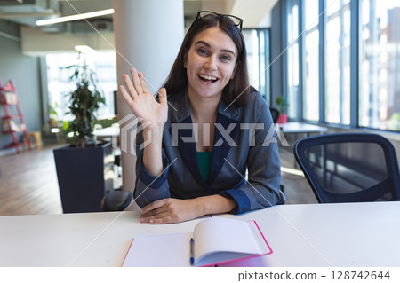 Woman sitting at white desk in open-plan office, waving with pink notebook and blue pen 128742644
