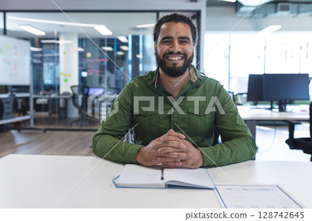 bi-racial young man sitting at desk in open-plan office, with notebook, monitors and sticky notes bi-racial young man sitting at desk in open-plan office, with notebook, monitors and sticky notes 128742645