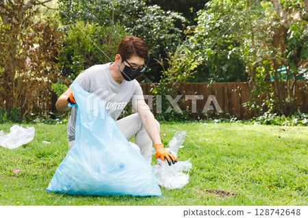 Asian man collecting plastic litter into blue bag on grassy park lawn, wearing orange gloves 128742648