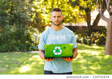 Male volunteer holding recycling bin and wearing orange gloves in sunlit garden promoting recycling 128742654
