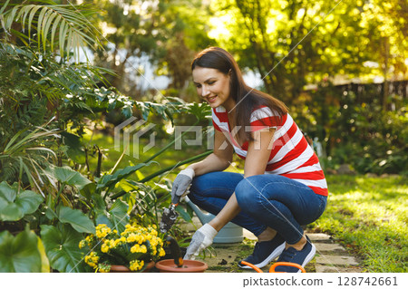 Woman crouching on path in backyard garden, planting yellow flowers with trowel and terracotta pots 128742661