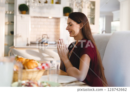 Woman sitting at wooden dining table in modern kitchen, praying with woven bread basket, copy space 128742662