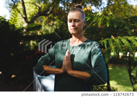 Senior woman practicing meditative yoga pose on wooden deck in backyard garden, with white couch Senior woman practicing meditative yoga pose on wooden deck in backyard garden, with white couch 128742673