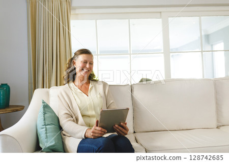 Woman sitting on cream-colored sofa in living room, holding tablet, smiling at screen, copy space Woman sitting on cream-colored sofa in living room, holding tablet, smiling at screen, copy space 128742685