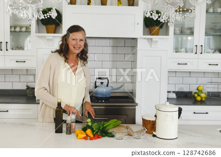 mature woman chopping vegetables on wooden cutting board in modern kitchen, with olive oil bottle 128742686