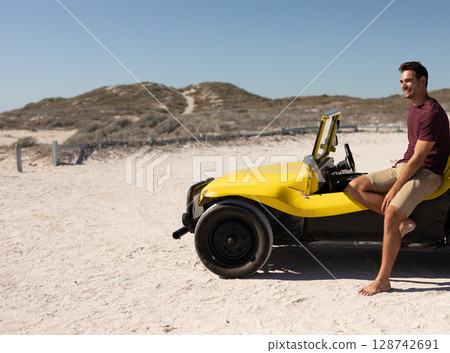 Man sitting barefoot on yellow dune buggy in coastal dunes with wooden posts, copy space Man sitting barefoot on yellow dune buggy in coastal dunes with wooden posts, copy space 128742691