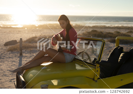Young woman sitting on hood of yellow dune buggy on beach at sunset, strumming acoustic guitar Young woman sitting on hood of yellow dune buggy on beach at sunset, strumming acoustic guitar 128742692