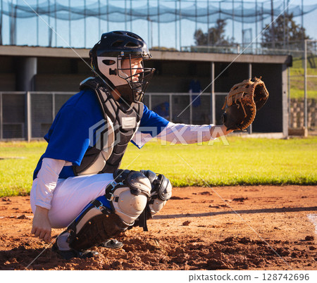 Young adult woman crouching on baseball infield dirt holding mitt near dugout fencing, copy space 128742696